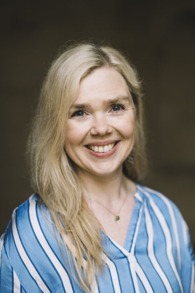 Eine Frau mit langen blonden Haaren, die eine blau-weiß gestreifte Bluse trägt, lächelt vor einem dunklen, verschwommenen Hintergrund in die Kamera.