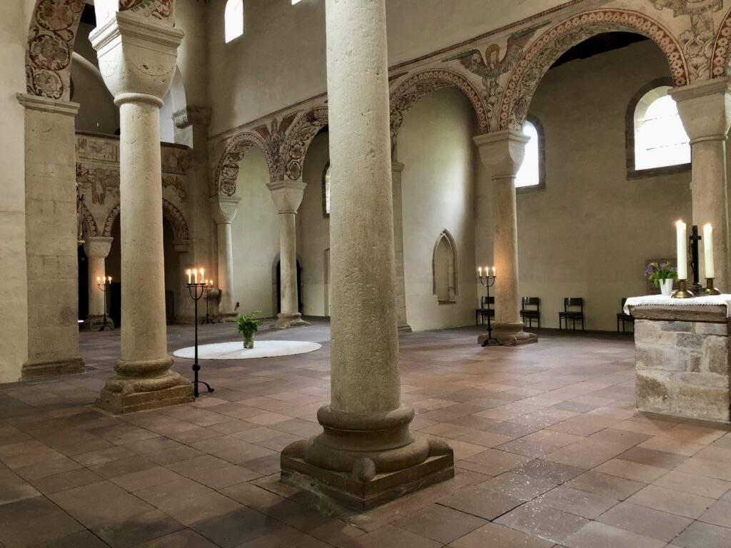 Das Innere einer alten Kirche mit Steinsäulen, verzierten Bögen, Kerzenständern und einem Altar mit brennenden Kerzen und Blumen. Durch die Bogenfenster fällt natürliches Licht ein.