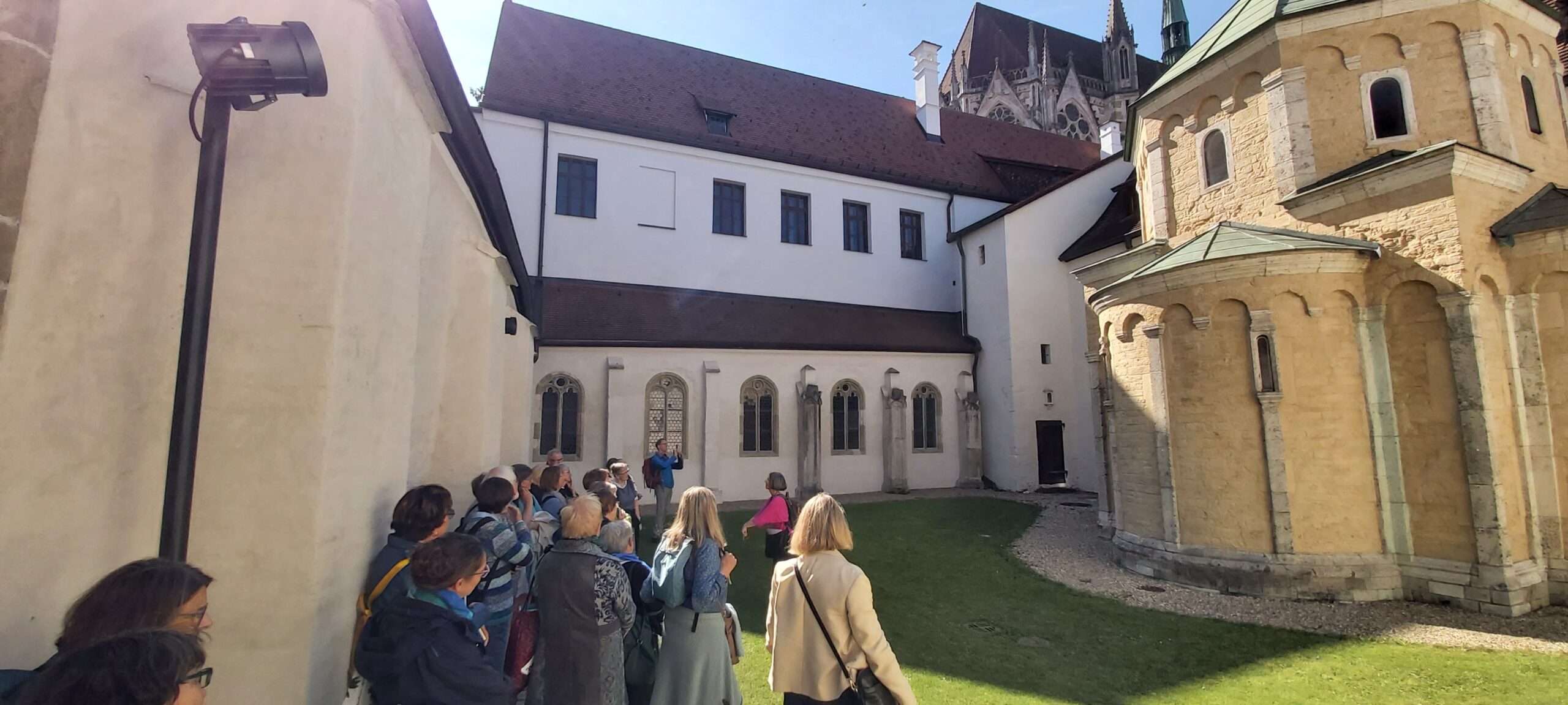 Eine Gruppe von Menschen spaziert durch einen sonnigen Innenhof mit historischen Steinhäusern, Bogenfenstern und einer Kirche mit Spitzdach im Hintergrund.