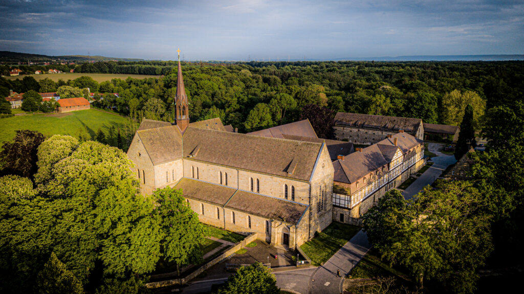 Luftaufnahme einer großen Steinkirche mit hohem Kirchturm, umgeben von grünen Bäumen und Feldern unter einem teilweise bewölkten Himmel.
