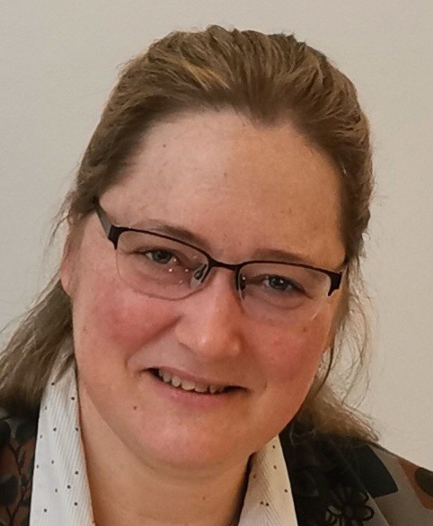 Eine Frau mit hellbraunen Haaren und Brille lächelt in die Kamera. Sie trägt ein weißes Hemd und eine Jacke mit dunklem Muster.