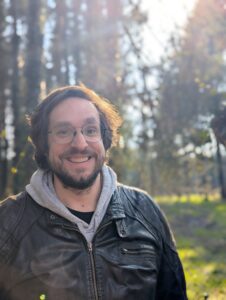 Ein Mann mit Brille und Bart lächelt draußen in einem sonnendurchfluteten Wald; er trägt eine schwarze Lederjacke über einem grauen Kapuzenpullover.
