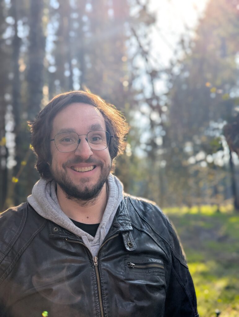 Ein Mann mit Brille und Bart lächelt draußen in einem sonnendurchfluteten Wald; er trägt eine schwarze Lederjacke über einem grauen Kapuzenpullover.