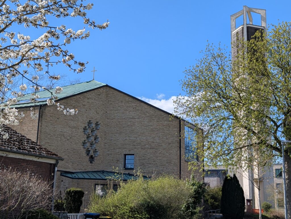 Eine moderne Kirche mit Backsteinfassade, hohem Glockenturm und begrüntem Dach, umgeben von blühenden Bäumen und viel Grün unter strahlend blauem Himmel.