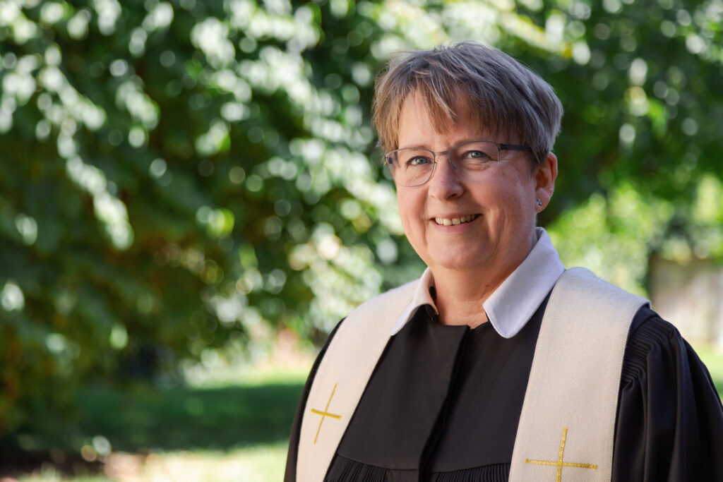 Eine lächelnde Person mit kurzen Haaren und Brille trägt eine Priesterrobe mit Stola und steht im Freien vor dem Hintergrund von Bäumen und Grünflächen.