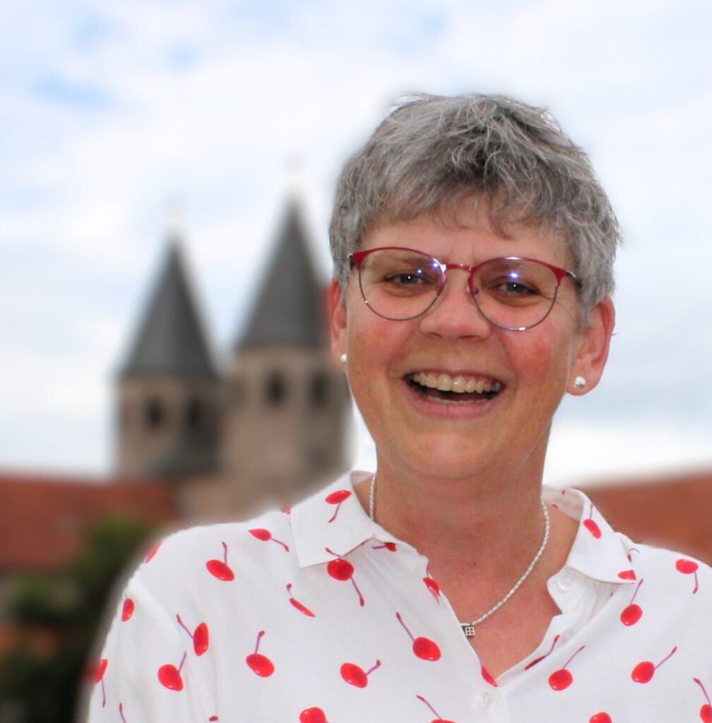 Lächelnde Frau mit kurzen grauen Haaren, Brille und weißem Hemd mit roten Kirschen, die im Freien steht, mit verschwommenen Kirchtürmen im Hintergrund.