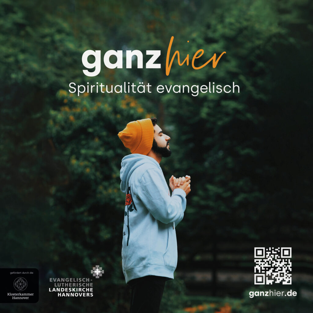 Eine bärtige Person mit einer leuchtend orangefarbenen Mütze und einem Kapuzenpulli steht im Freien, die Augen geschlossen und die Hände gefaltet, und scheint zu beten. Der Text lautet: „ganz hier. Spiritualität evangelisch.“.