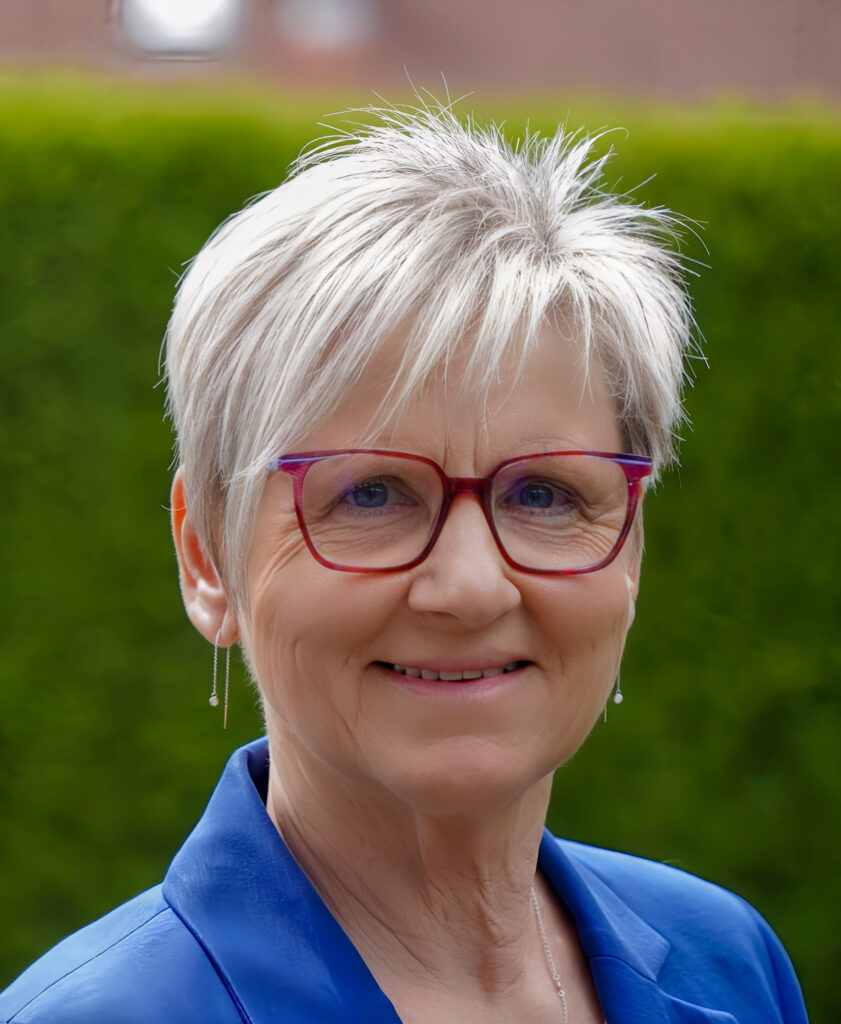 Eine lächelnde ältere Frau mit kurzen grauen Haaren, die eine rote Brille und einen blauen Blazer trägt und draußen vor einer grünen Hecke steht.