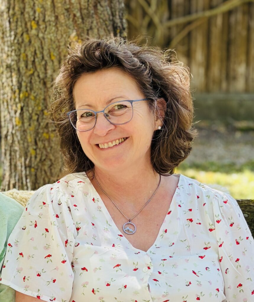 Eine Frau mit lockigem braunem Haar und Brille sitzt draußen auf einer Bank und lächelt; sie trägt eine weiße Bluse mit Blumenmuster und eine silberne Halskette.