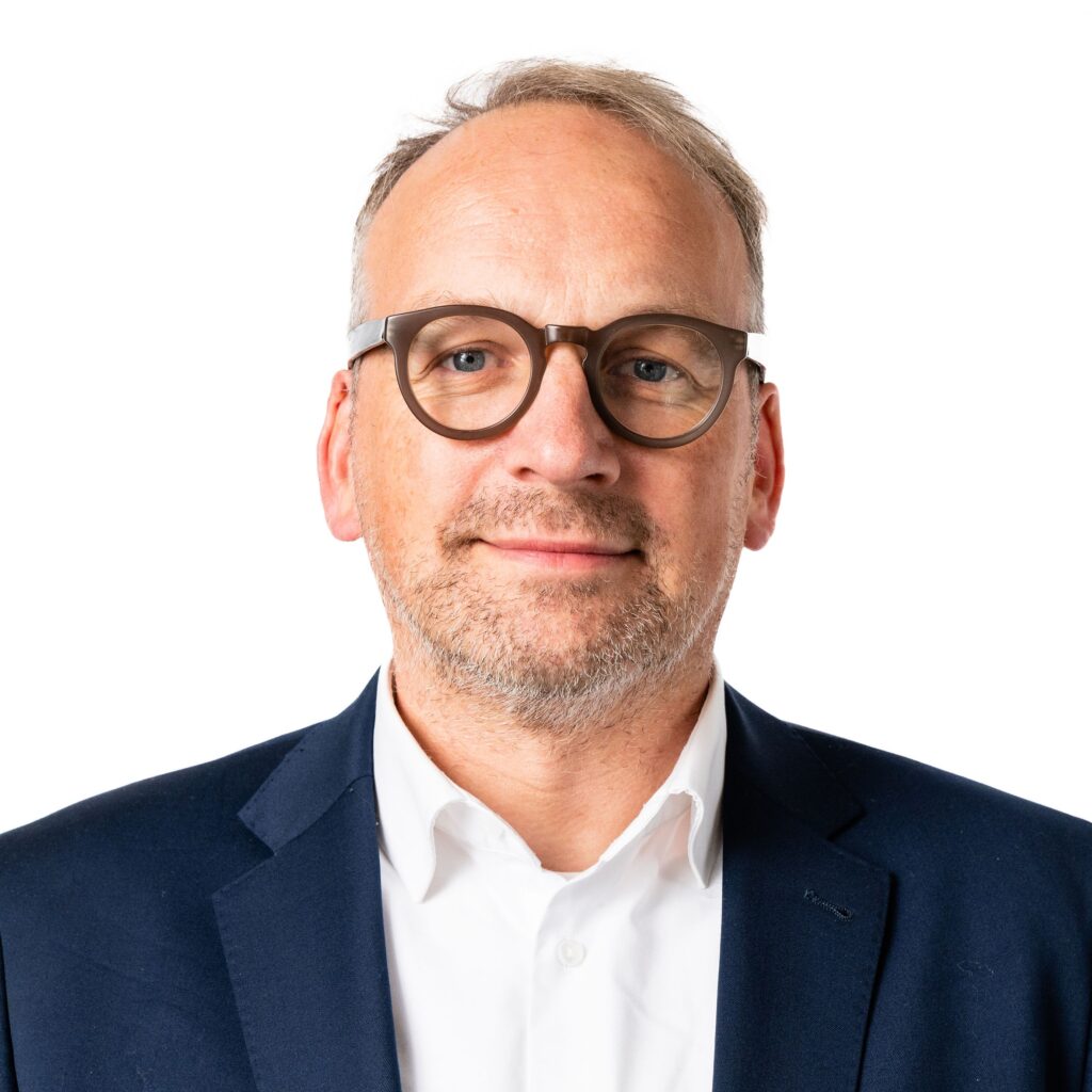 Ein Mann mit kurzen, hellen Haaren, Brille und Bart, der einen marineblauen Anzug und ein weißes Hemd trägt und vor einem schlichten weißen Hintergrund steht.