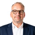 Ein Mann mit kurzen, hellen Haaren, Brille und Bart, der einen marineblauen Anzug und ein weißes Hemd trägt und vor einem schlichten weißen Hintergrund steht.