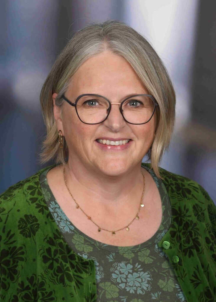 Lächelnde Frau mit kurzen grauen Haaren und Brille, die goldene Ohrringe und ein grünes Oberteil mit Blumenmuster trägt, vor einem sanft unscharfen Hintergrund.