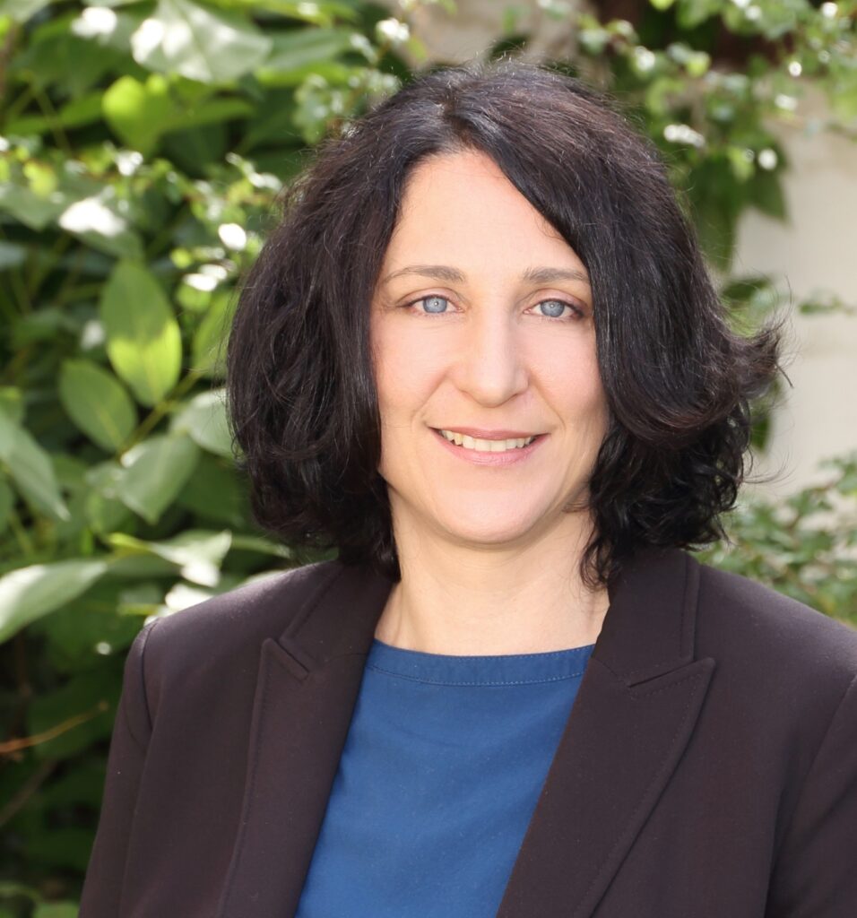 Eine Frau mit schulterlangem dunklem Haar, die einen dunklen Blazer und ein blaues Oberteil trägt, lächelt im Freien vor grünen Blattpflanzen.