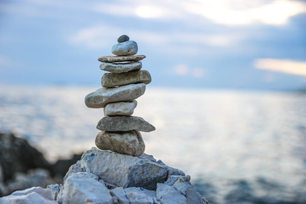 Ein Stapel glatter Steine balanciert auf felsigem Untergrund am Wasser, im Hintergrund sind der Himmel und das Meer nur verschwommen zu erkennen.