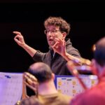Ein Dirigent mit lockigem Haar und Brille leitet die Musiker bei einer Probe an; im Vordergrund sind Notenständer und Blechblasinstrumente zu sehen.
