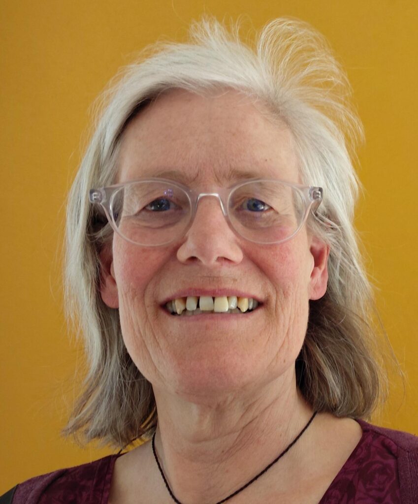 Eine lächelnde ältere Frau mit grauen Haaren, einer klaren Brille und einem weinroten Oberteil steht vor einem senfgelben Hintergrund.