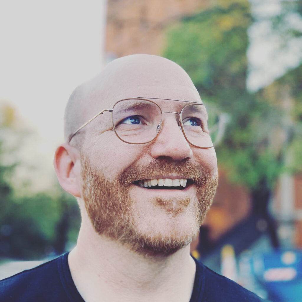 Ein lächelnder Mann mit Brille und Bart blickt im Freien nach oben, im Hintergrund sind Bäume und ein Backsteingebäude verschwommen zu sehen.