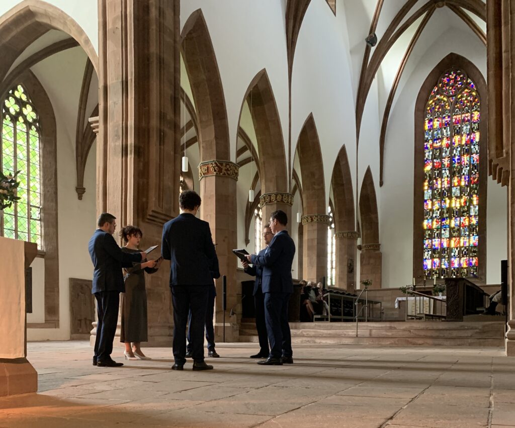 Eine kleine Gruppe von Menschen in festlicher Kleidung steht während einer Zeremonie in einer Steinkirche mit Gewölbedecken und Buntglasfenstern im Kreis.