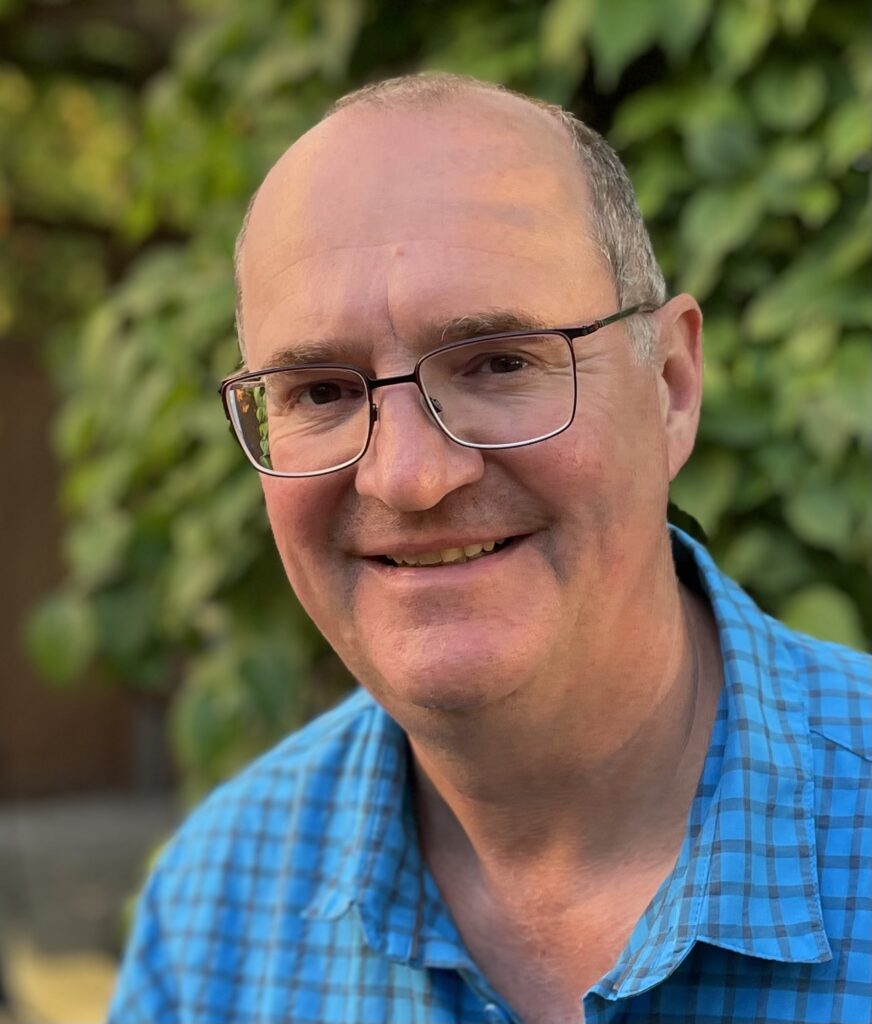 Ein Mann mittleren Alters mit Brille und einem blau karierten Hemd lächelt vor einem grünen, begrünten Hintergrund in die Kamera.